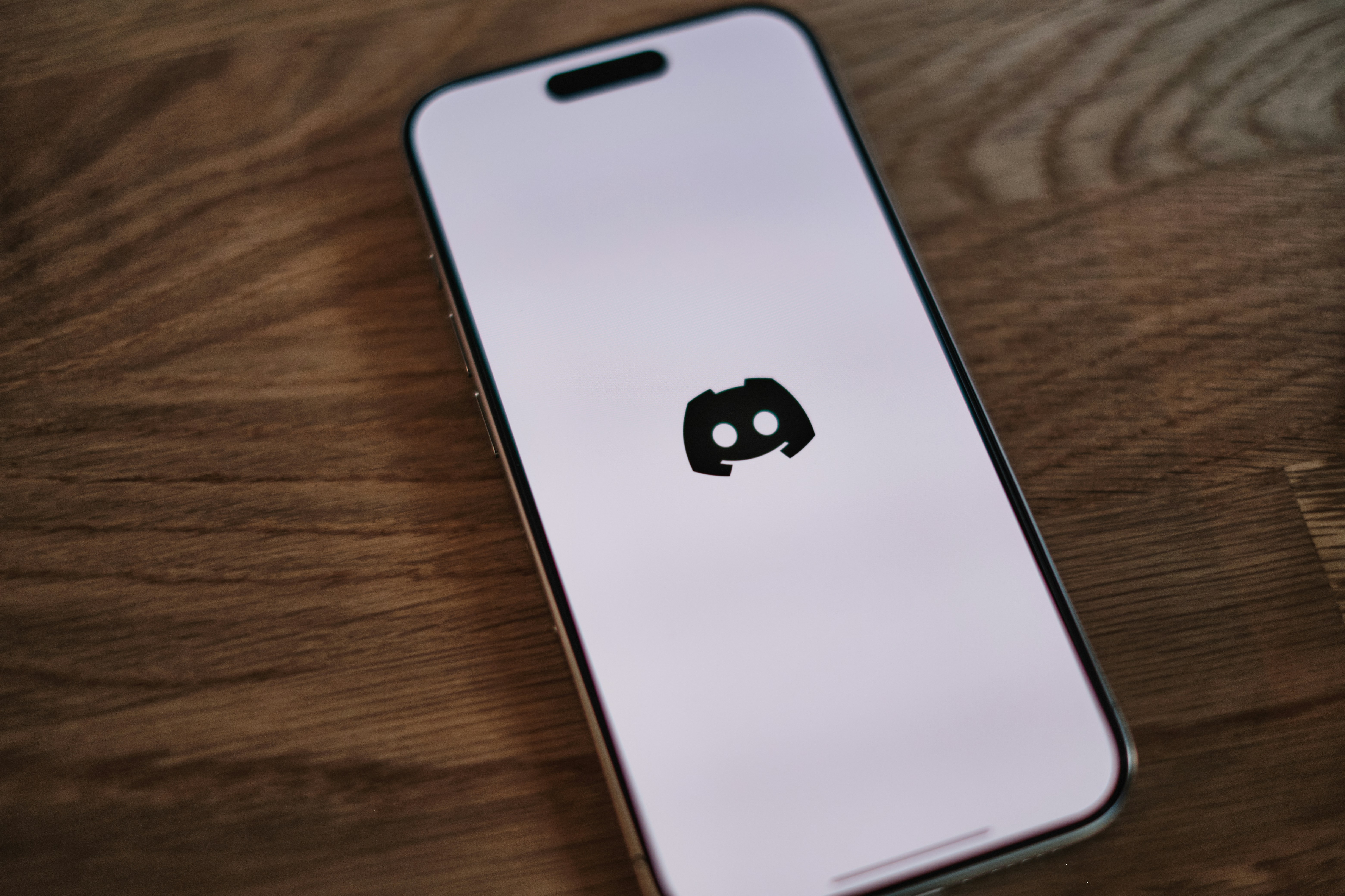 A smartphone resting on a wooden surface displays the black Discord logo on a white loading screen.
