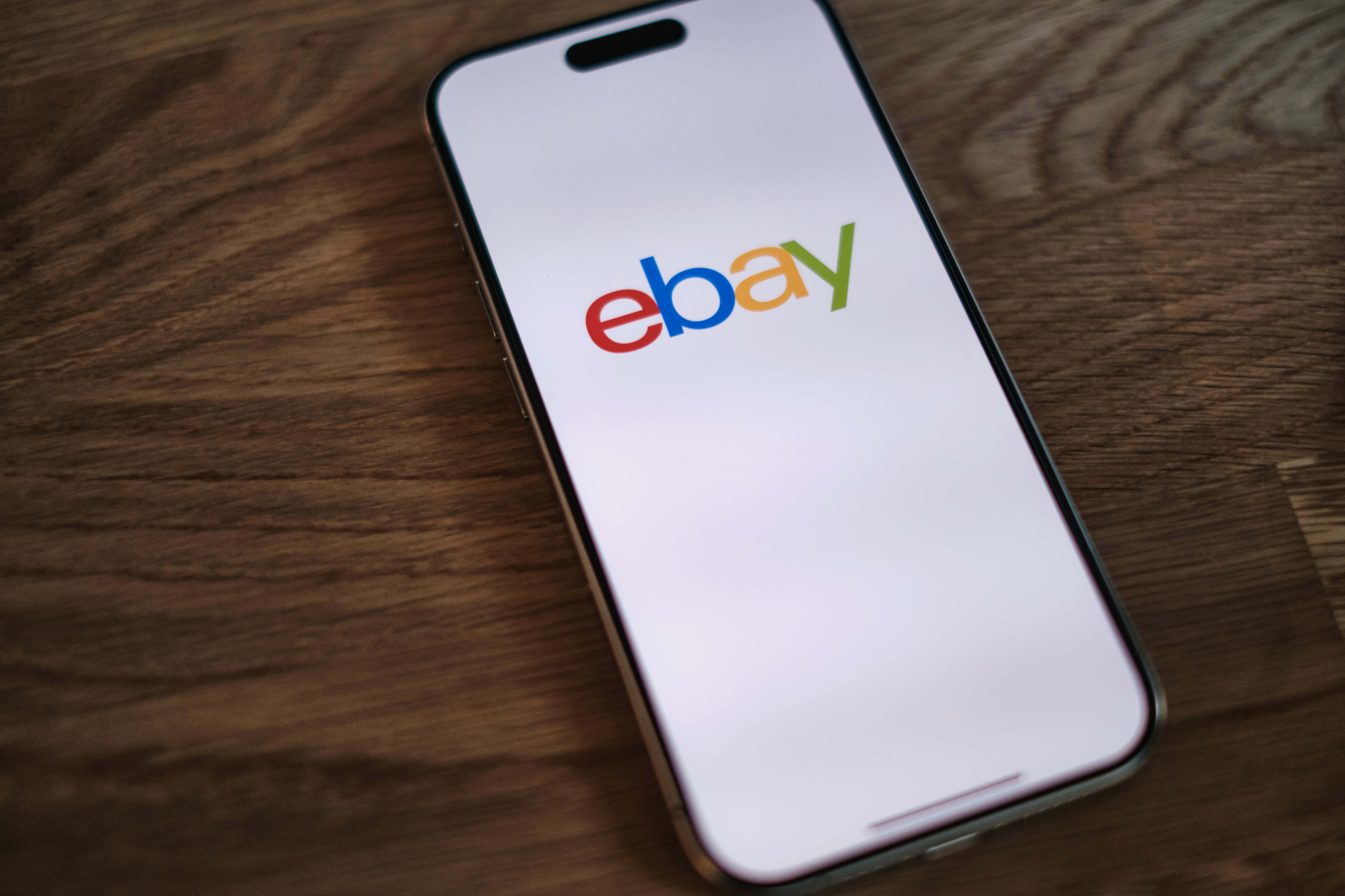 A smartphone displays the colorful eBay logo on its screen while resting on a dark wooden surface.