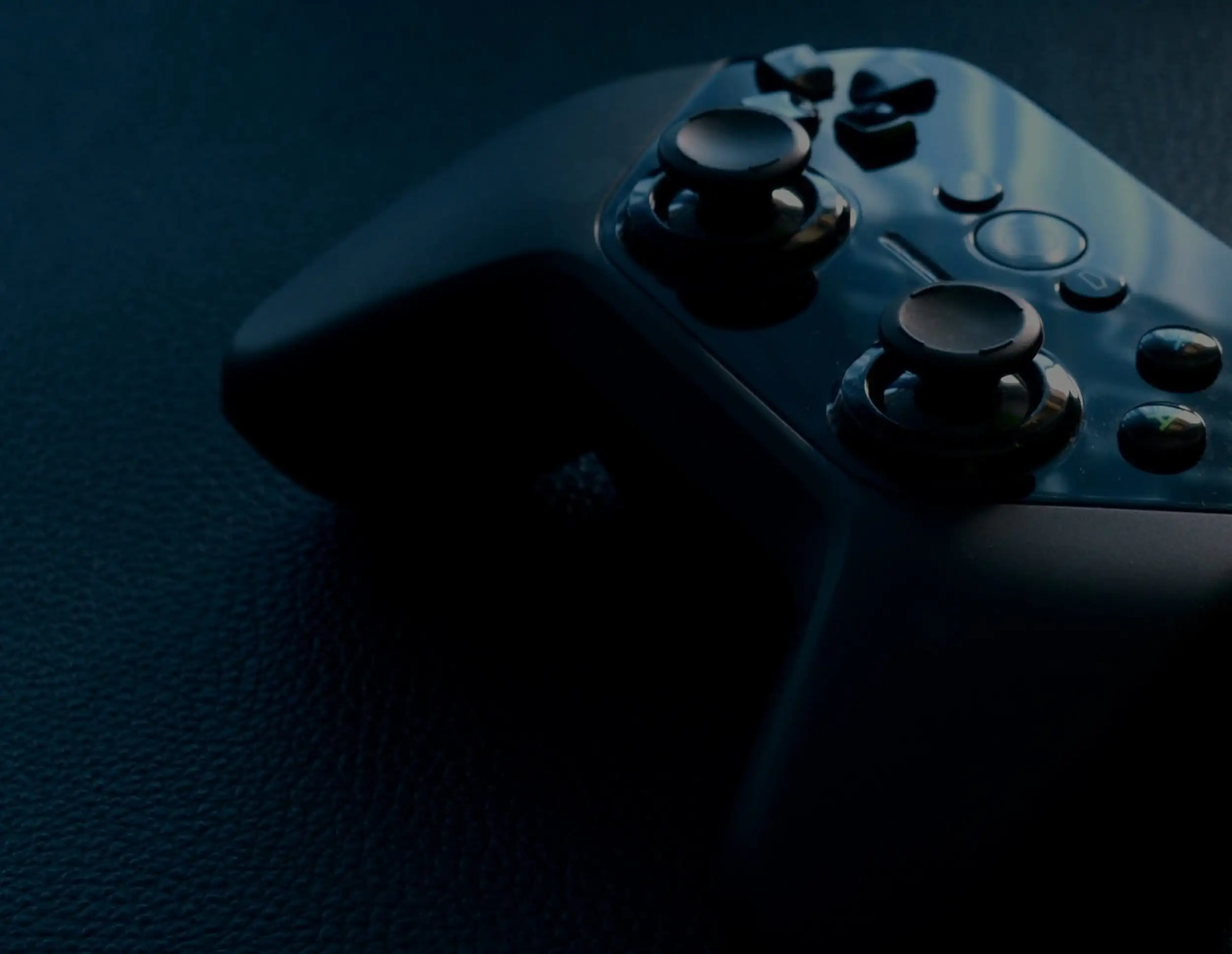 A black video game controller with dual joysticks and several buttons rests on a dark, textured surface. The lighting is low, casting subtle reflections on the controllers glossy surface.