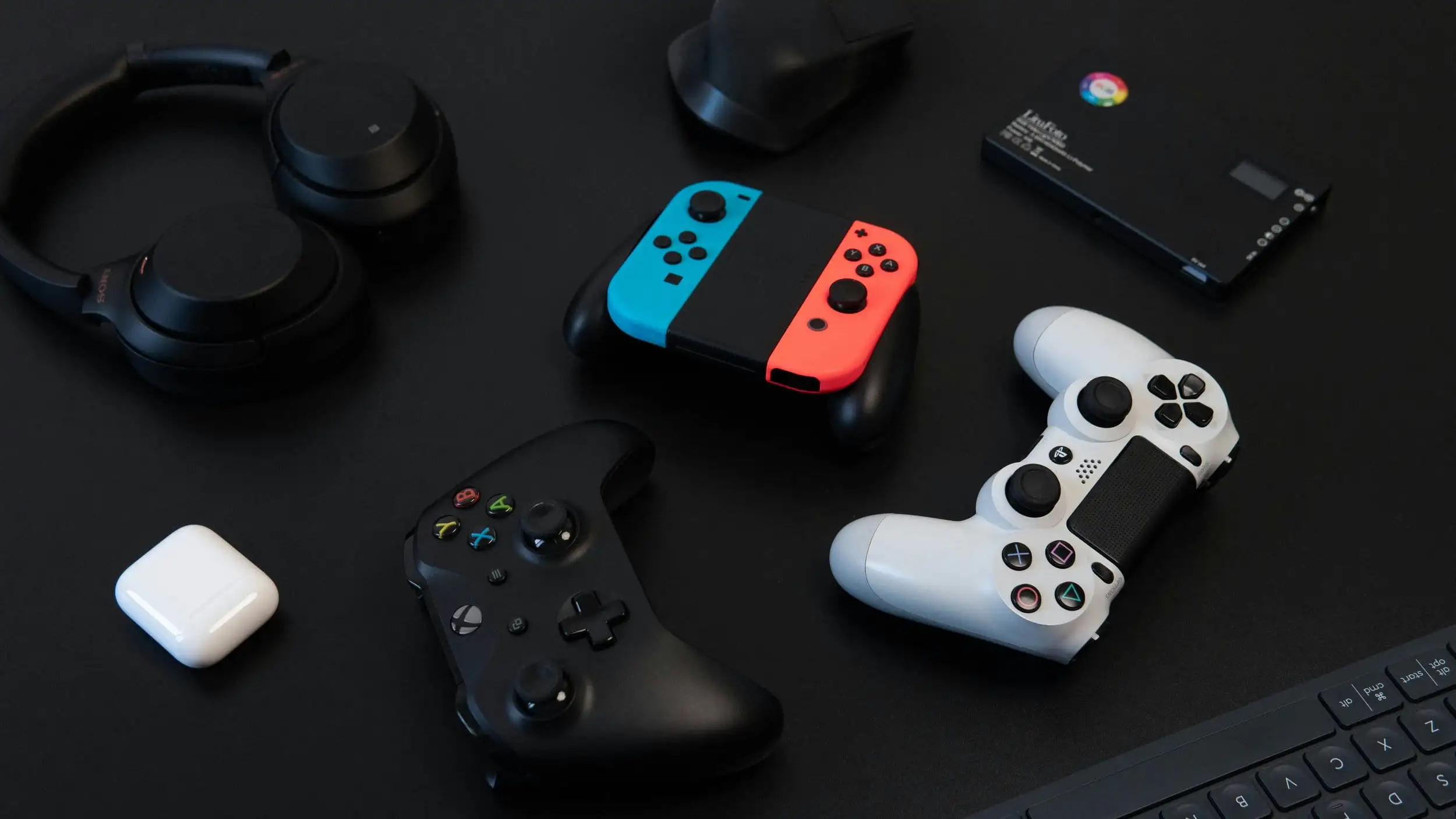 A selection of gaming and tech devices on a black surface, including black wireless headphones, a red and blue handheld game controller, a black controller, a white controller, a pair of white earbuds, and a small black box with a multicolored logo.