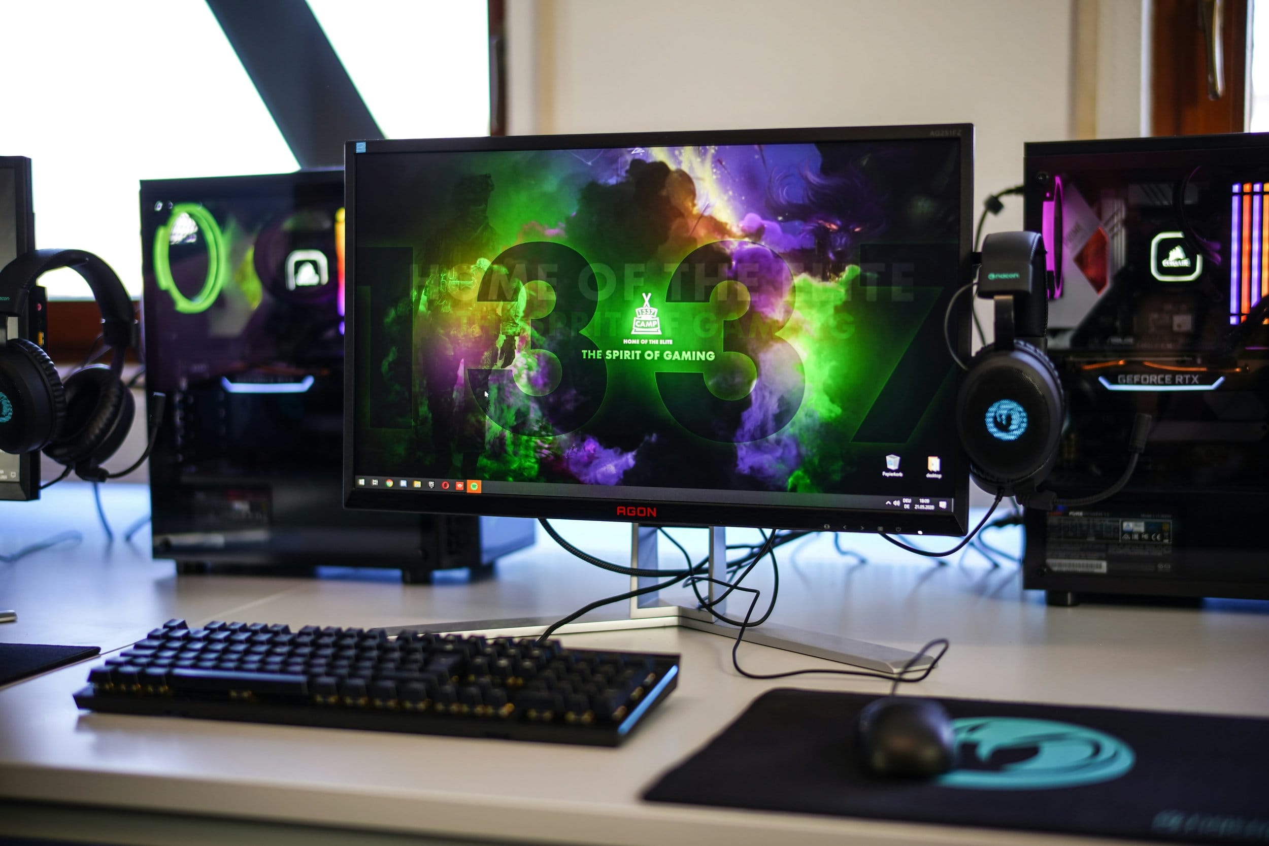 Dual custom-built PCs with RGB lighting, AGON monitor, headset, and mechanical keyboard on a gaming desk.