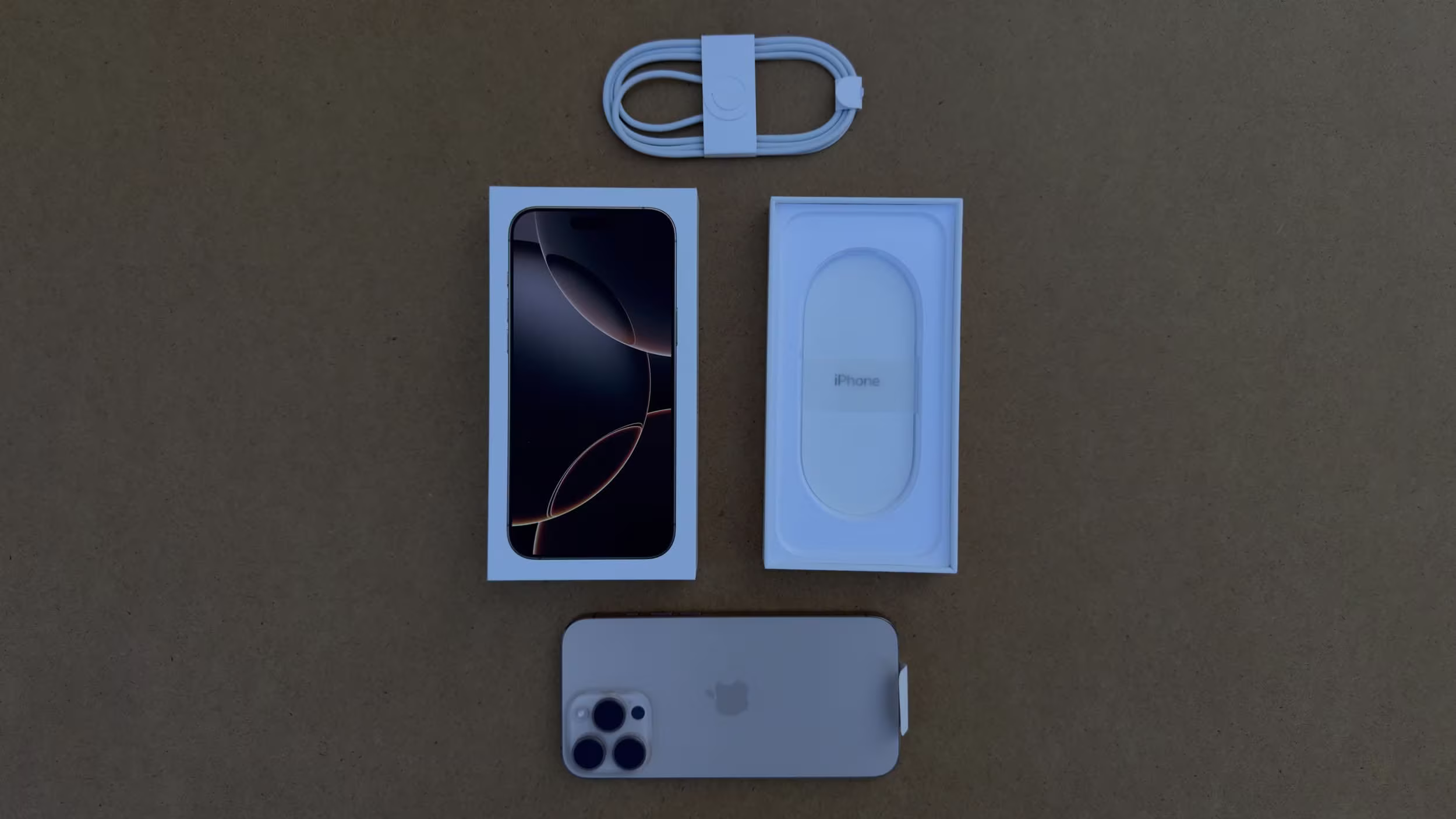 Top-down view of the full iPhone 16 unboxing contents including the phone, box, USB-C cable, and documentation neatly laid out on a brown surface.