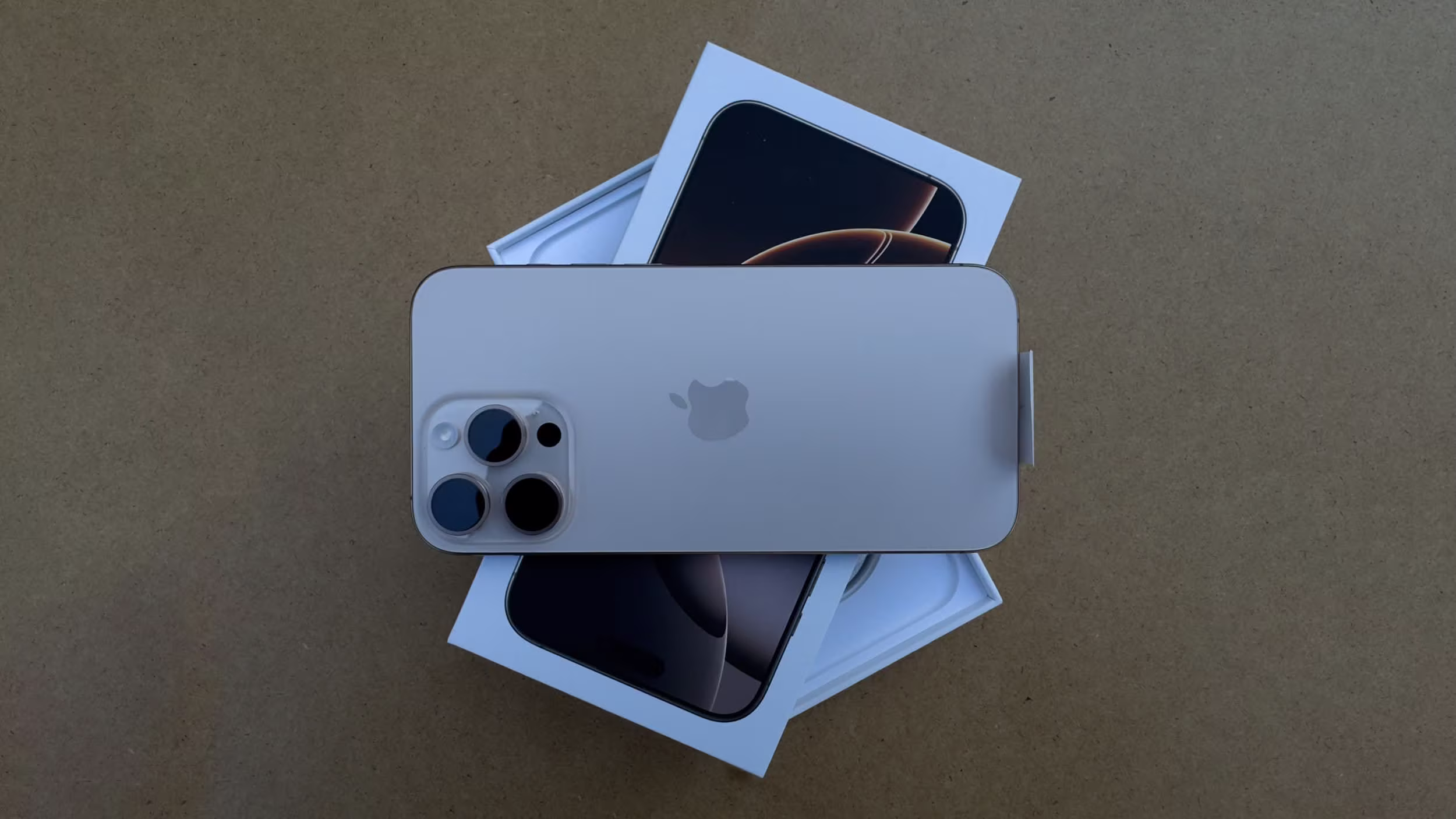 A top-down view of the iPhone 16 resting on its open box, showing the back of the phone with its triple camera layout and Apple logo.