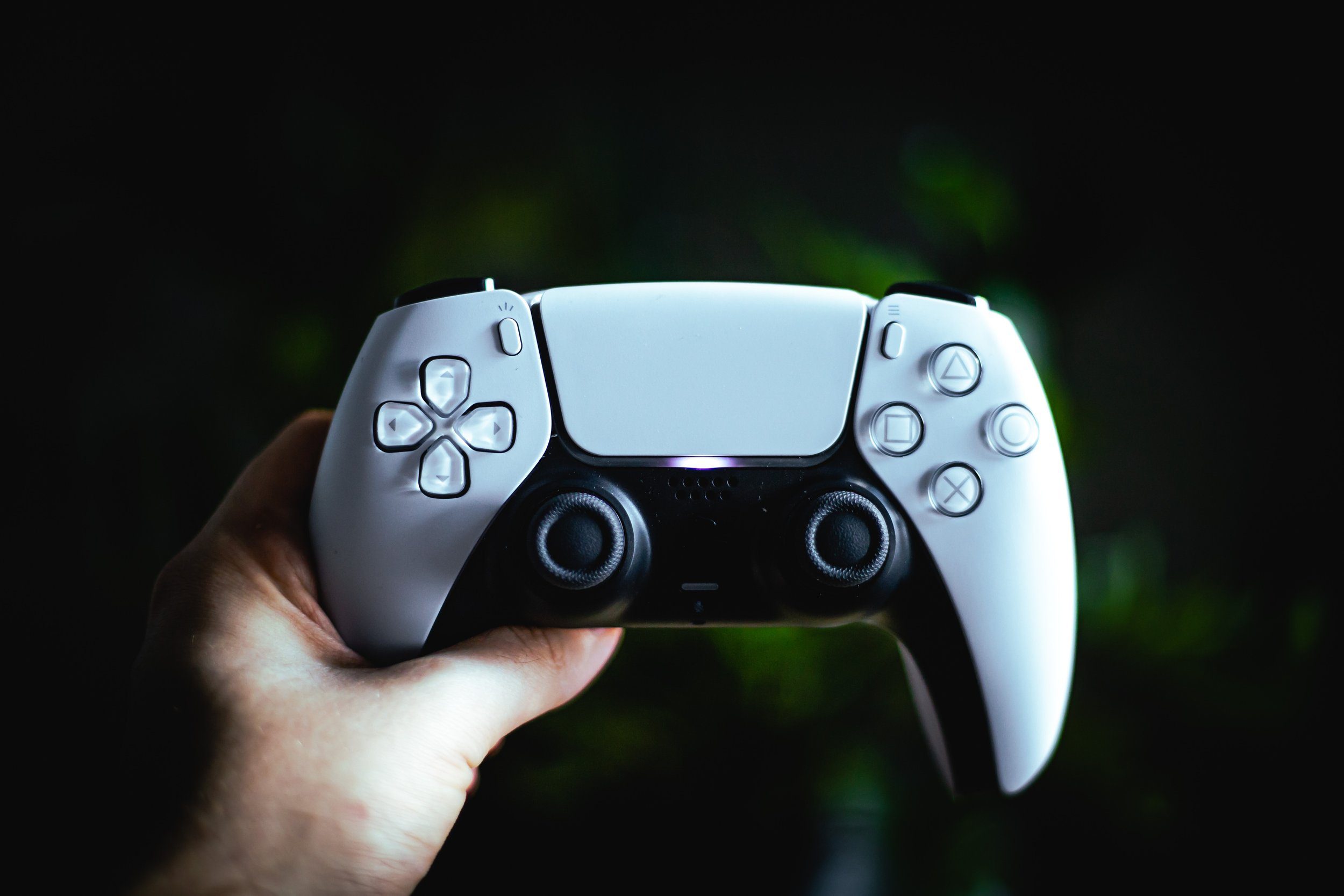 Close-up of a hand holding a white PS5 DualSense controller against a dark background with green tones