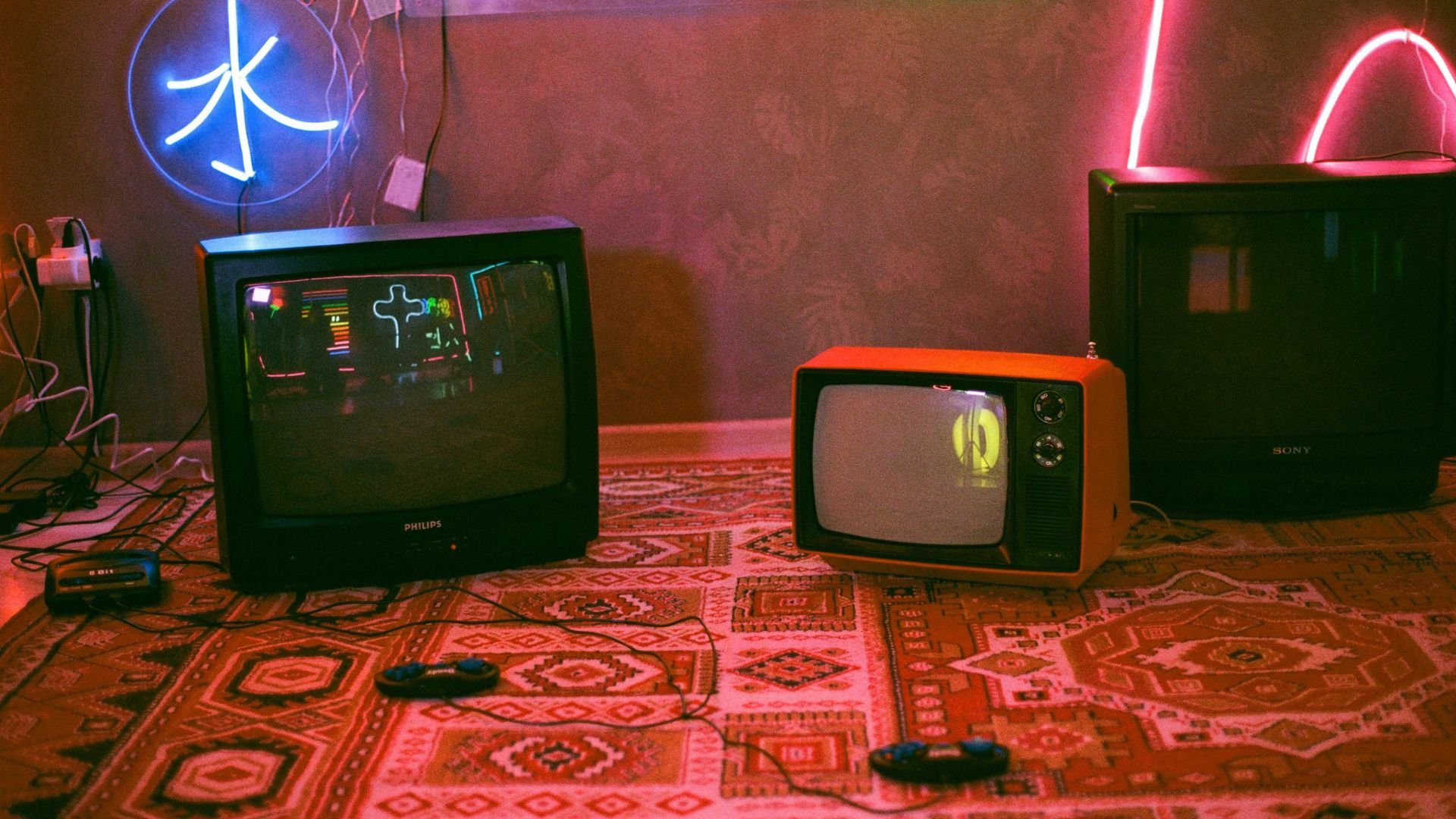 Three retro CRT TVs sitting on a patterned carpet with wired gaming controllers nearby and neon lights on the wall.